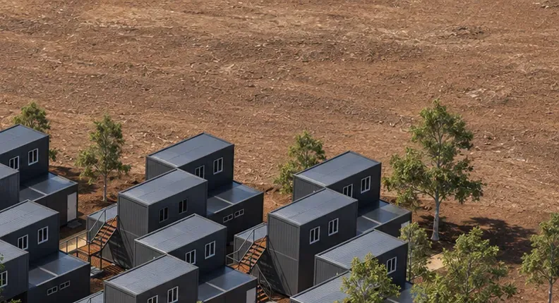 Terreno de tierra seca con árboles dispersos junto a módulos prefabricados grises